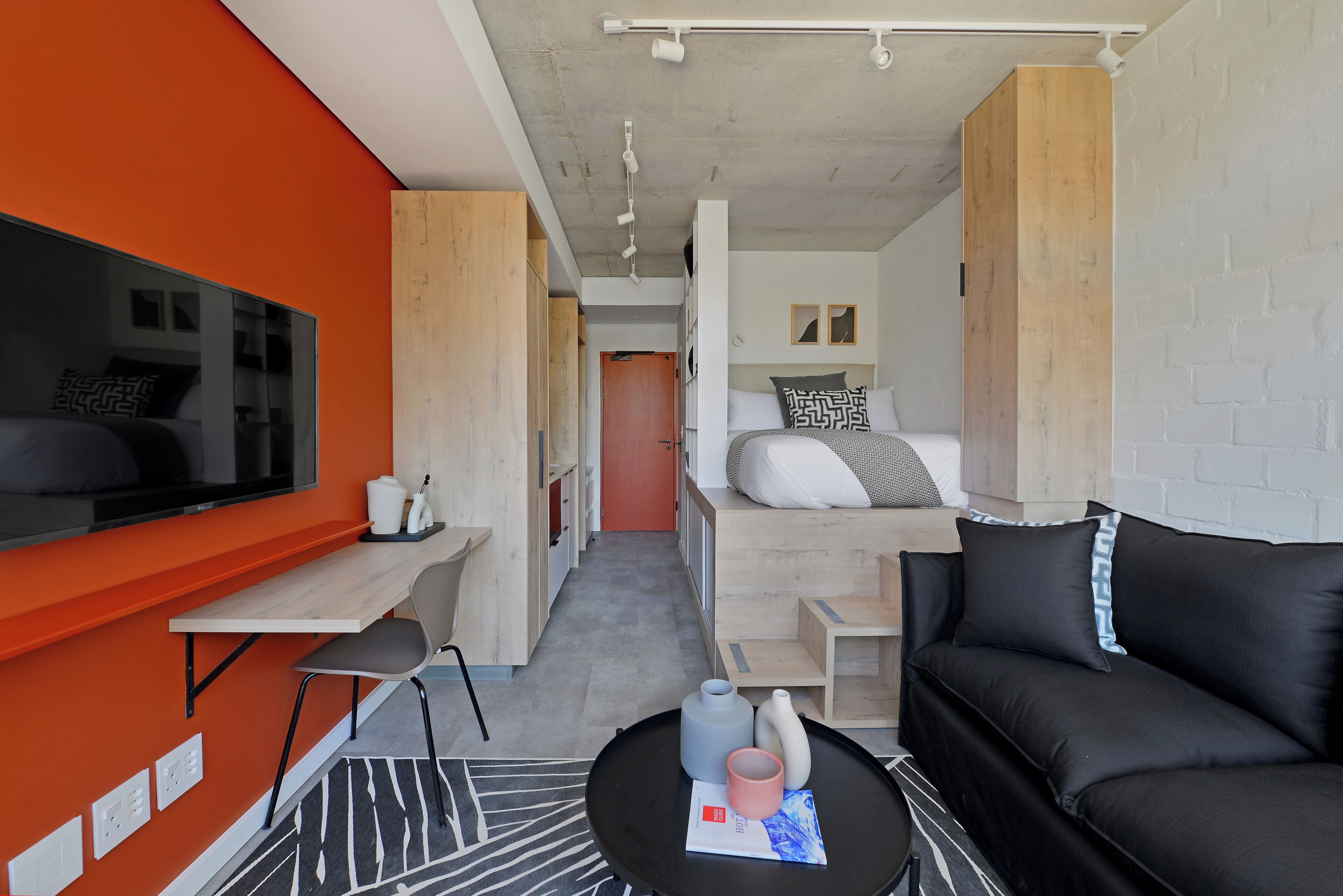 Una habitación compacta y moderna con cama, sofá negro, escritorio con silla y televisor.