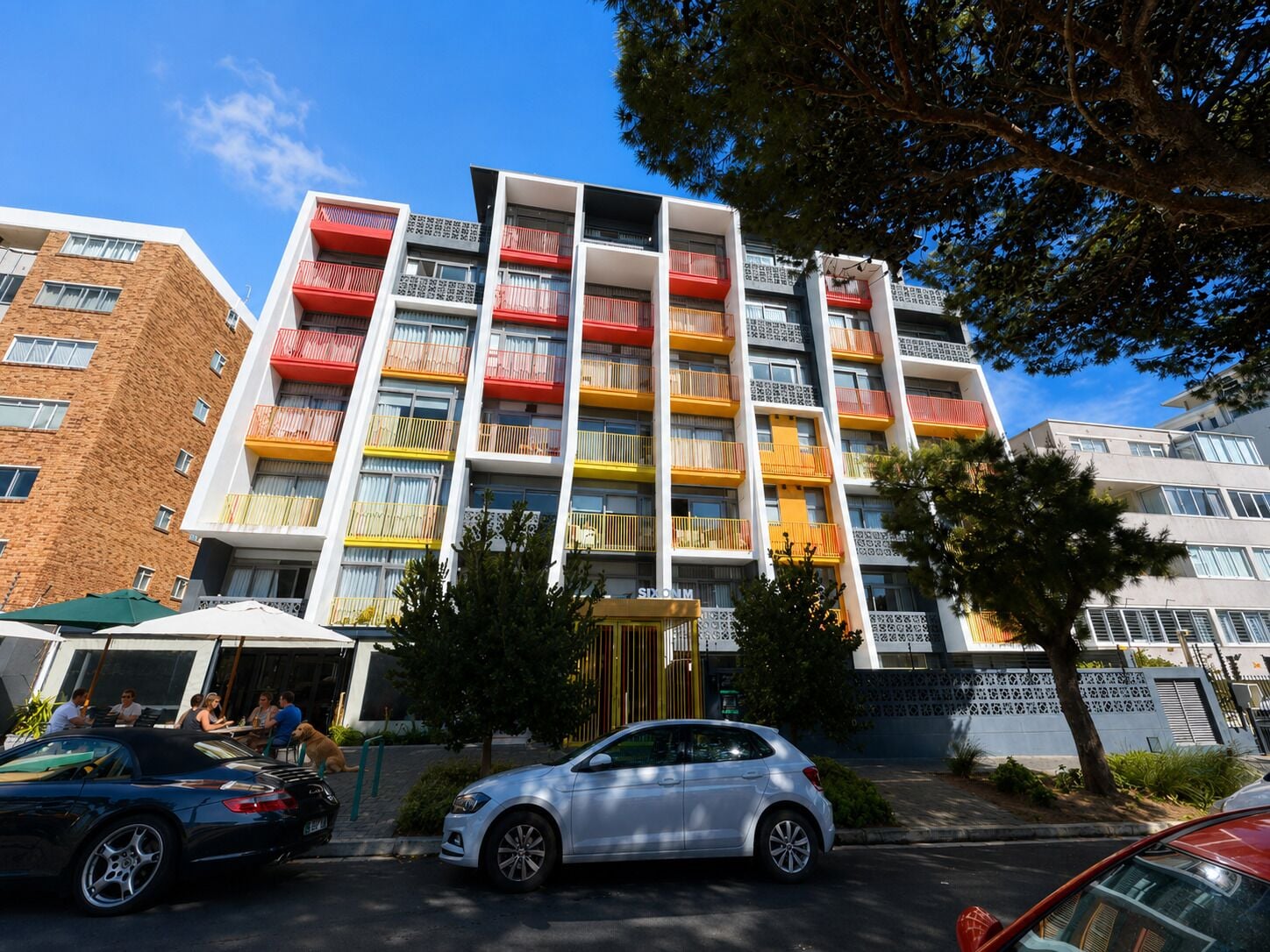 A multi-story building with balconies, a gated entrance, and a parking area with cars.