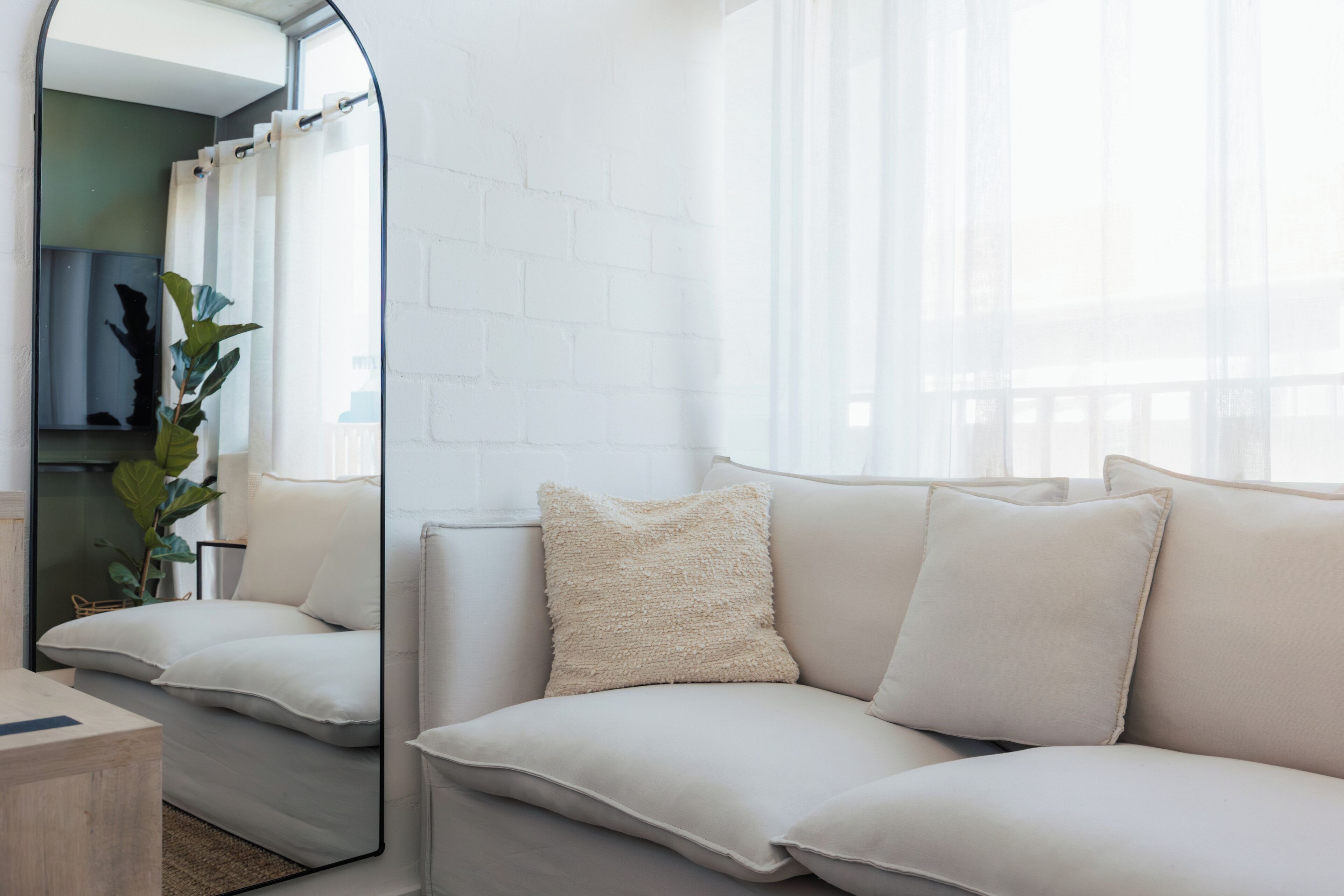 A modern living room with a white brick wall, a large mirror, a beige sofa with cushions, and a potted plant.