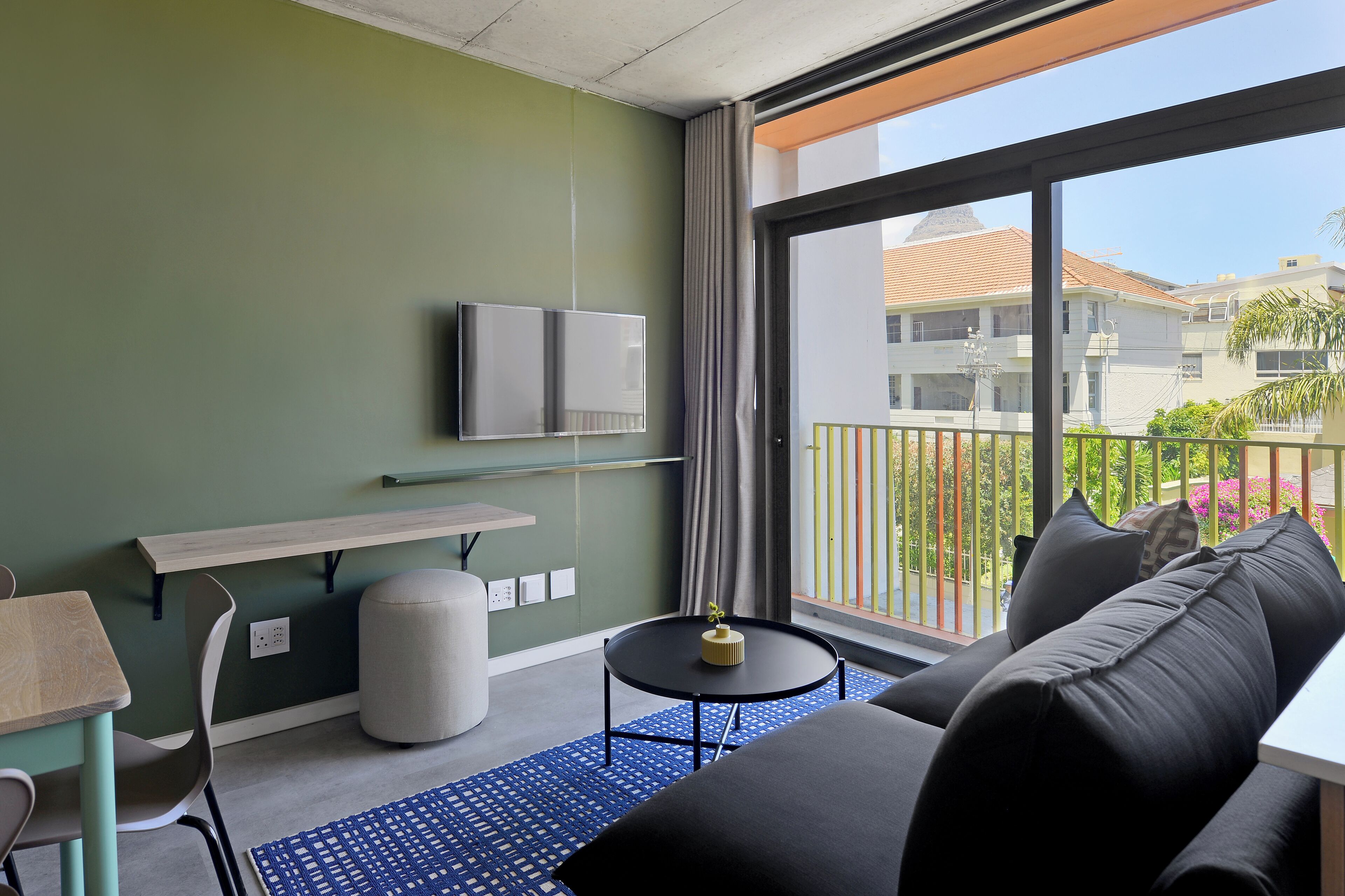Une chambre d’hôtel moderne, dotée d’un canapé, d’une petite table ronde, d’un téléviseur fixé au mur et d’un balcon offrant une vue sur la ville.