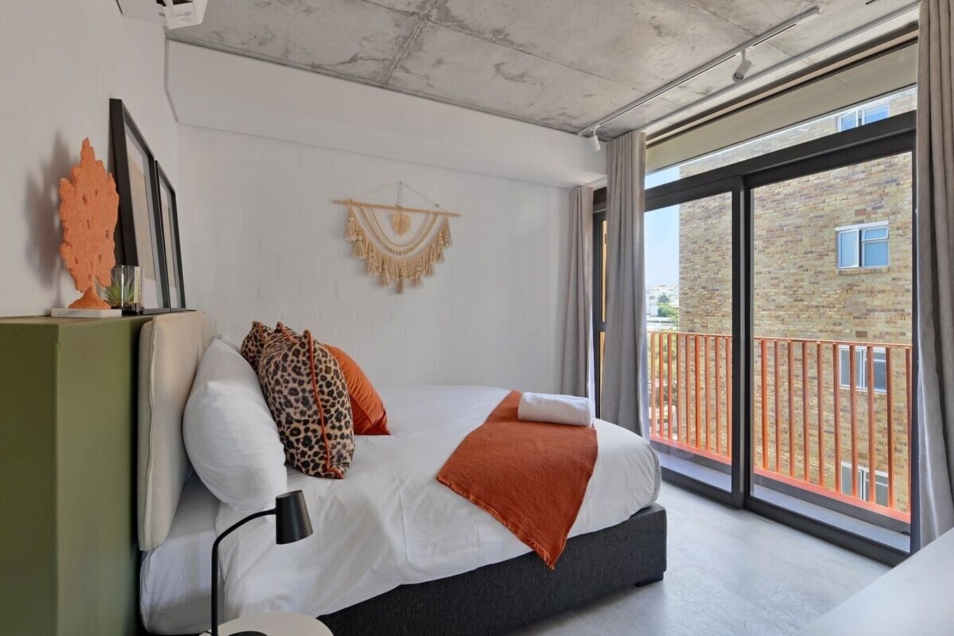 A modern bedroom with a large bed, a side table, a wall-mounted macrame piece, and a view of a brick building through a sliding glass door.