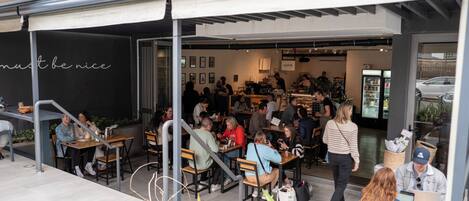 Ein Straßencafé mit Tischen und Stühlen, Personen beim Essen, und ein Schild mit der Aufschrift 'must be nice'.