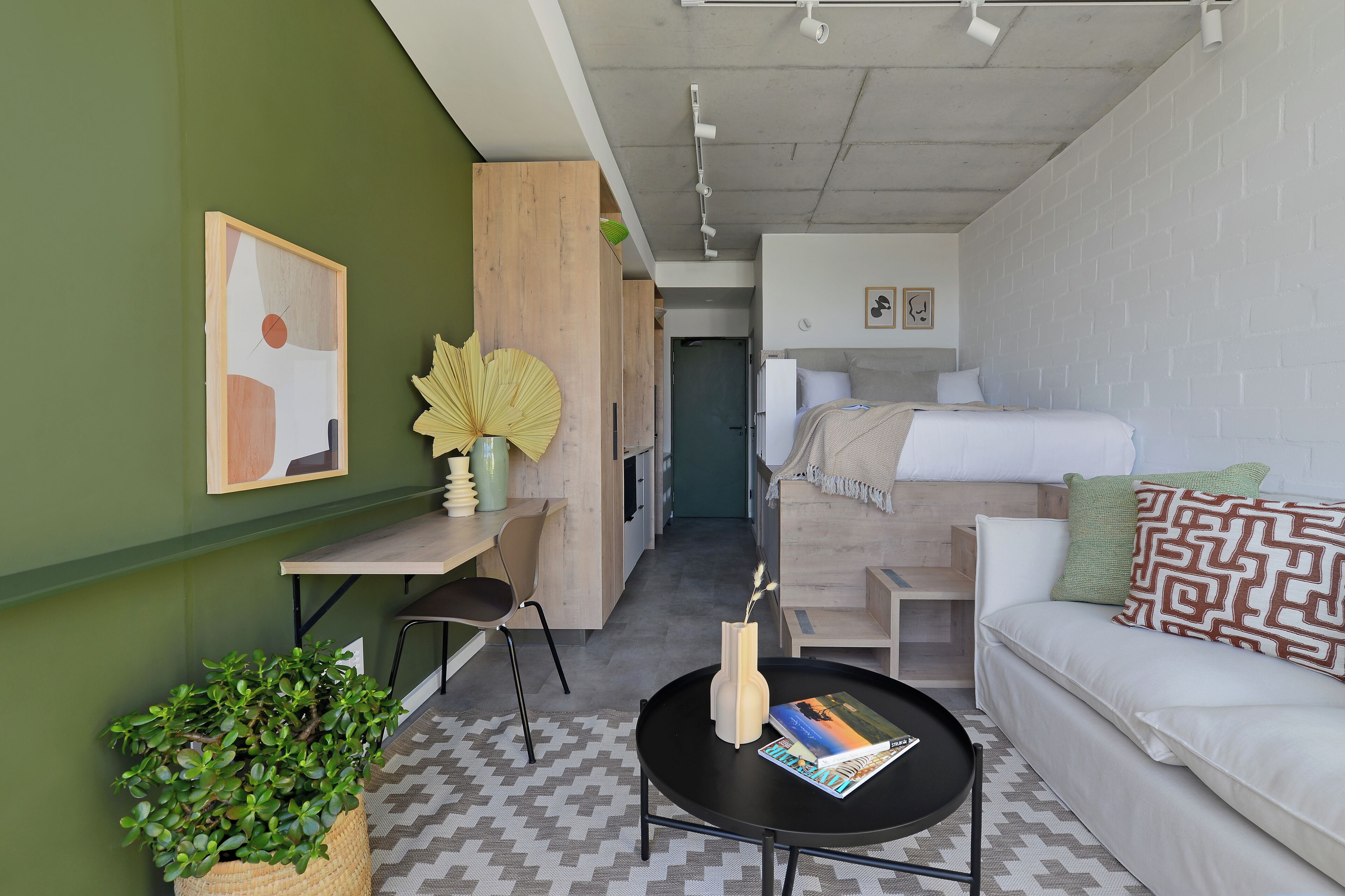 A compact hotel room with a bed, a sofa, a small table, a chair, a potted plant, and a wall-mounted artwork.
