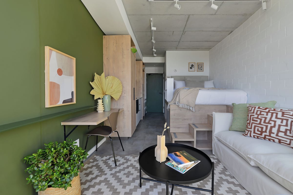 Ett kompakt hotellrum med en säng, en soffa, ett litet bord, en stol, en krukväxt och ett konstverk som hänger på väggen.