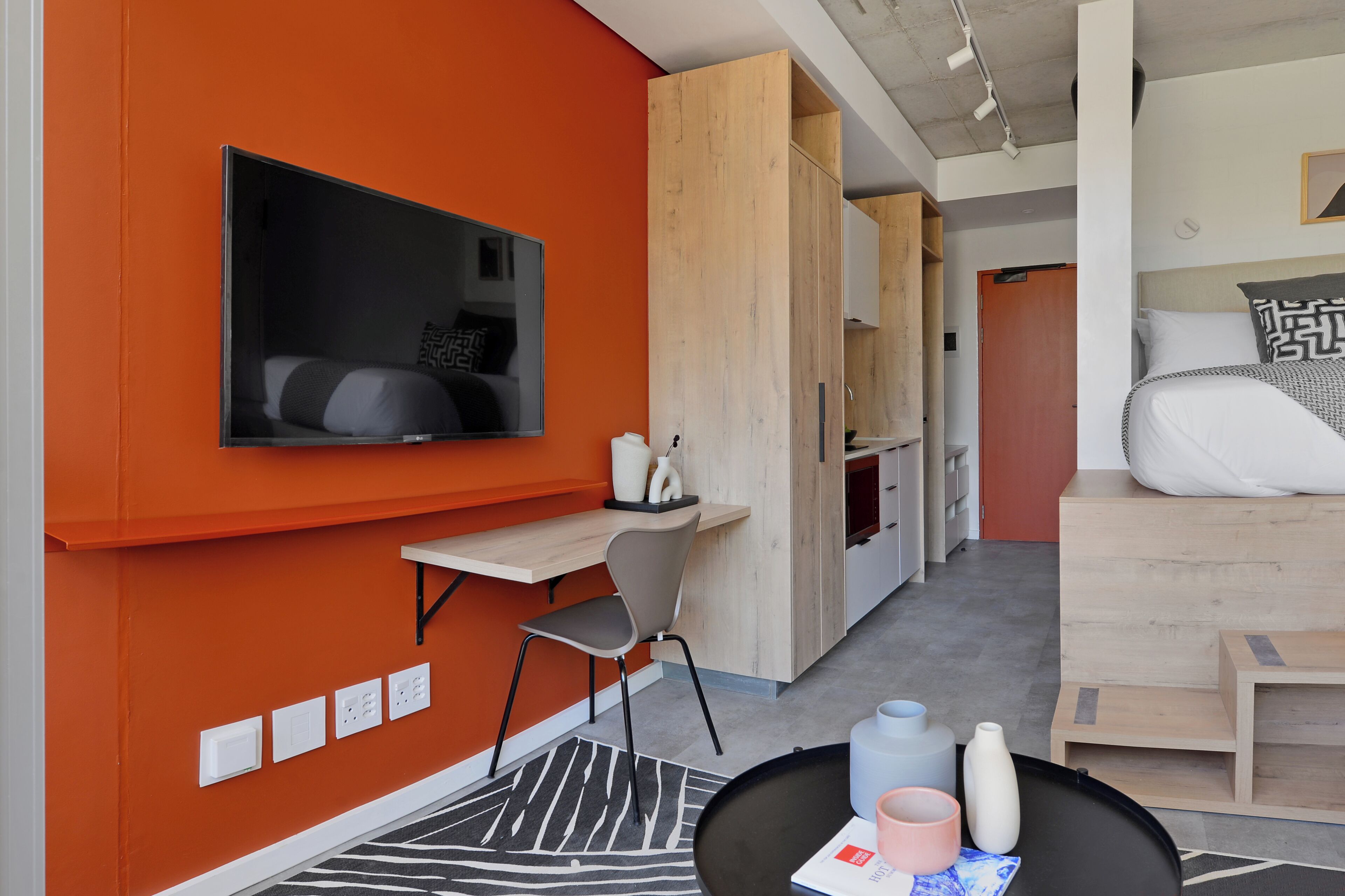 A modern hotel room with a TV mounted on the wall, a wooden desk, a chair, a sofa, and a small table with a magazine.