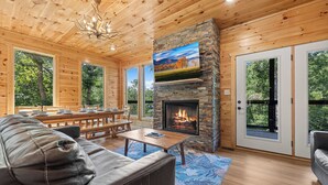 A cozy cabin interior with a stone fireplace, wooden ceiling, and large windows offering a view of the forest. - New Listing! Indoor pool, hot tub, theater, bocce, billiards, & firepit. (Sevierville)