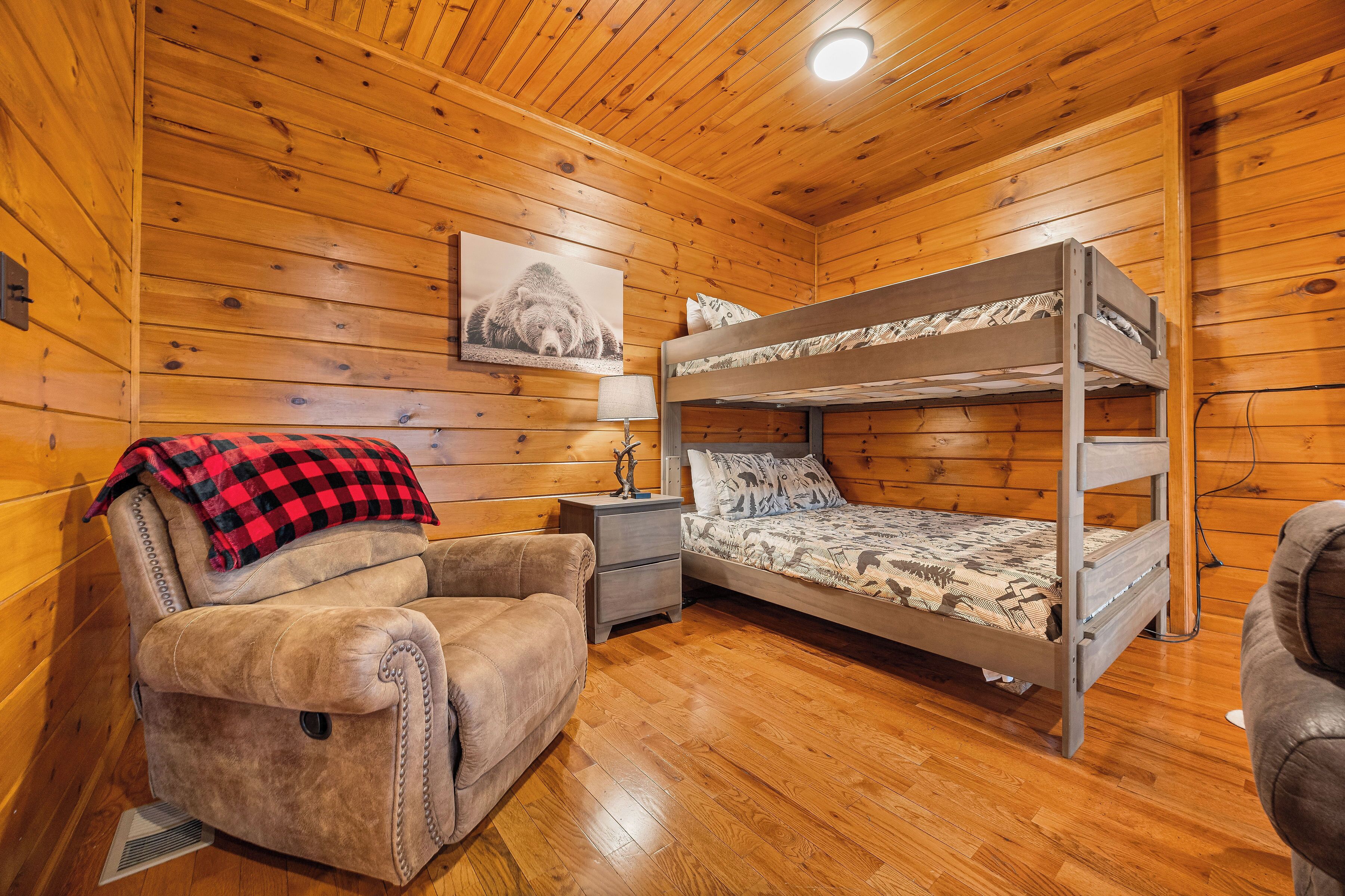 Una habitación cálida tipo cabaña con una litera, un sillón acolchado, una mesita de noche con lámpara y un cuadro en la pared.