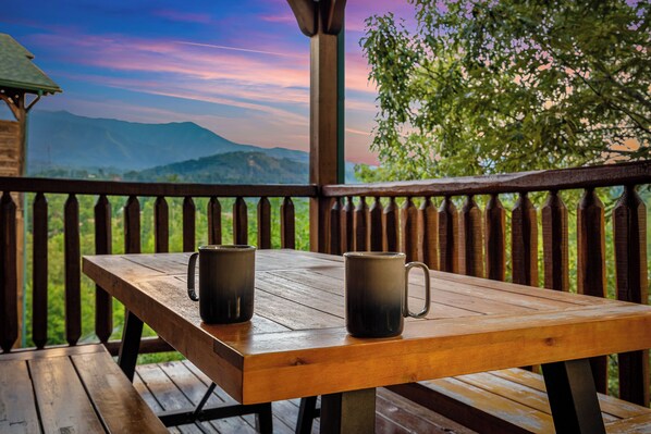 A wooden table with two dark mugs on a wooden deck with a railing, overlooking a mountainous landscape.