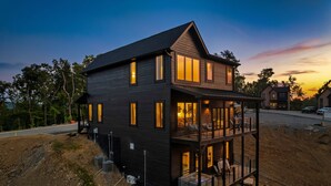 A two-story house with multiple windows and a balcony, surrounded by trees and a road. - Brand New Modern Luxe 6 Bed w/ Arcade Games, Indoor Pool, Great Views! (Sevierville)