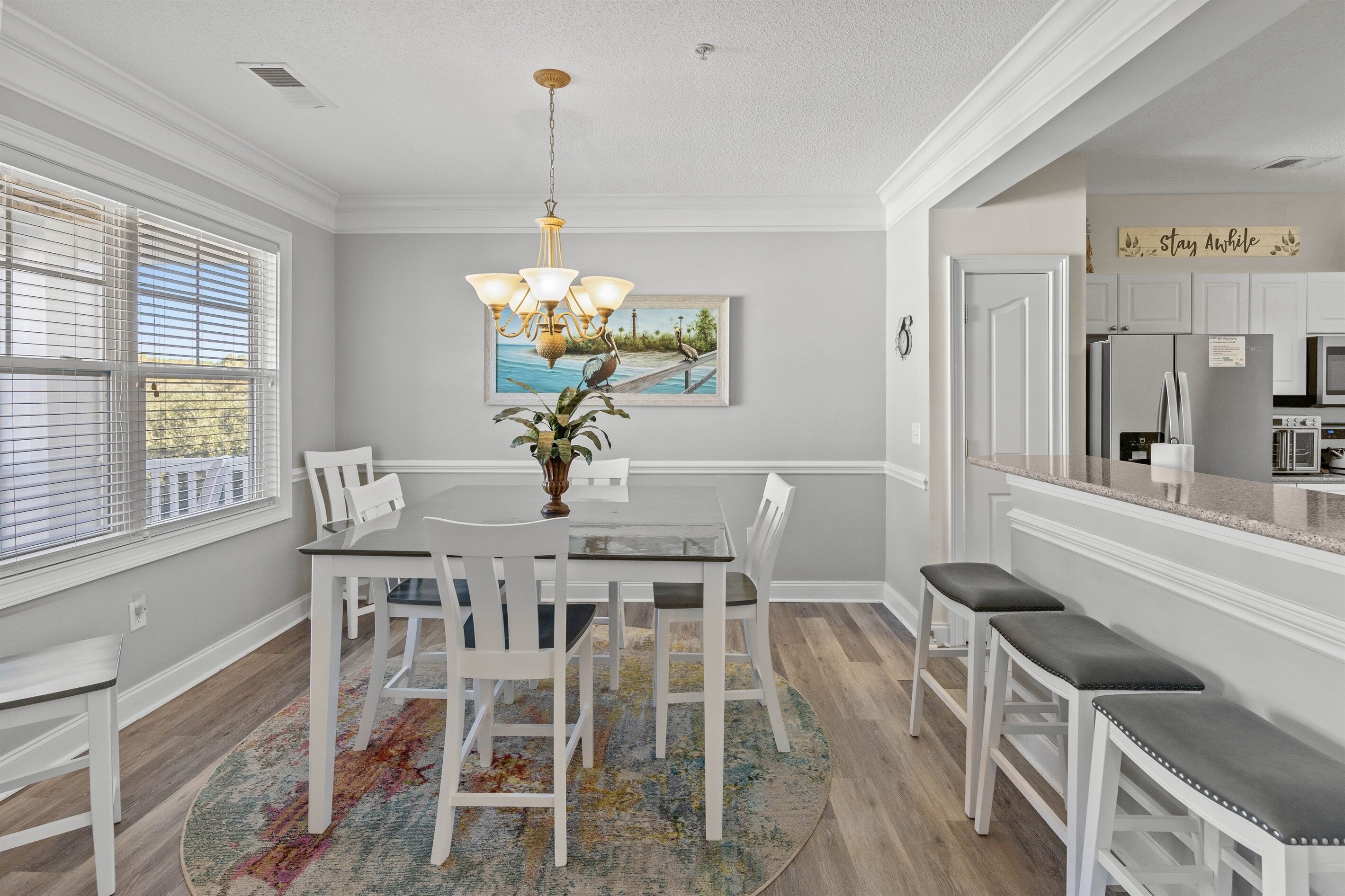 A dining area with a round table, white chairs, and a chandelier. There is a kitchen area with white cabinets and a countertop.