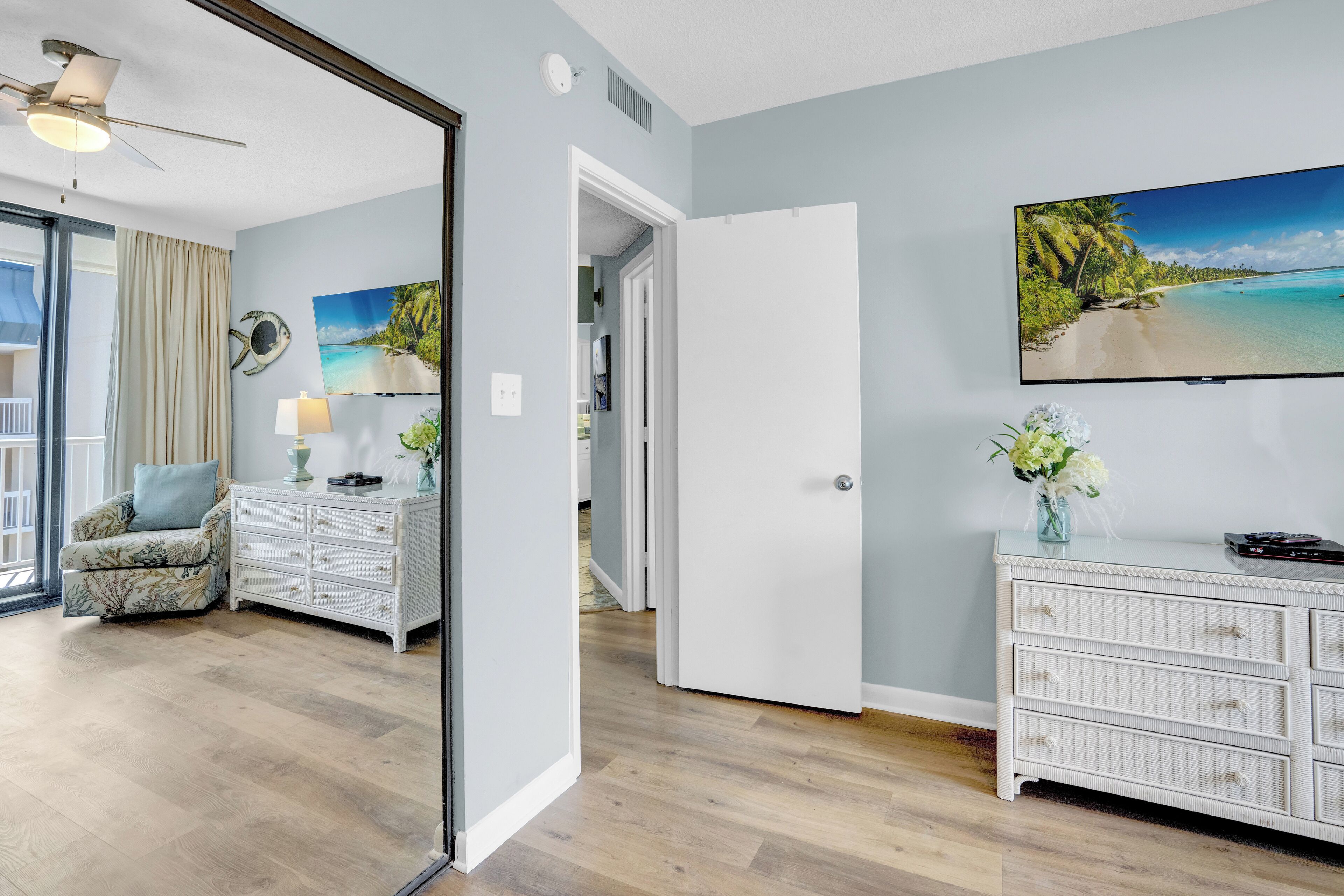 Une pièce avec un plancher en bois, une commode blanche, une chaise, un ventilateur de plafond et un tableau représentant une scène de plage.