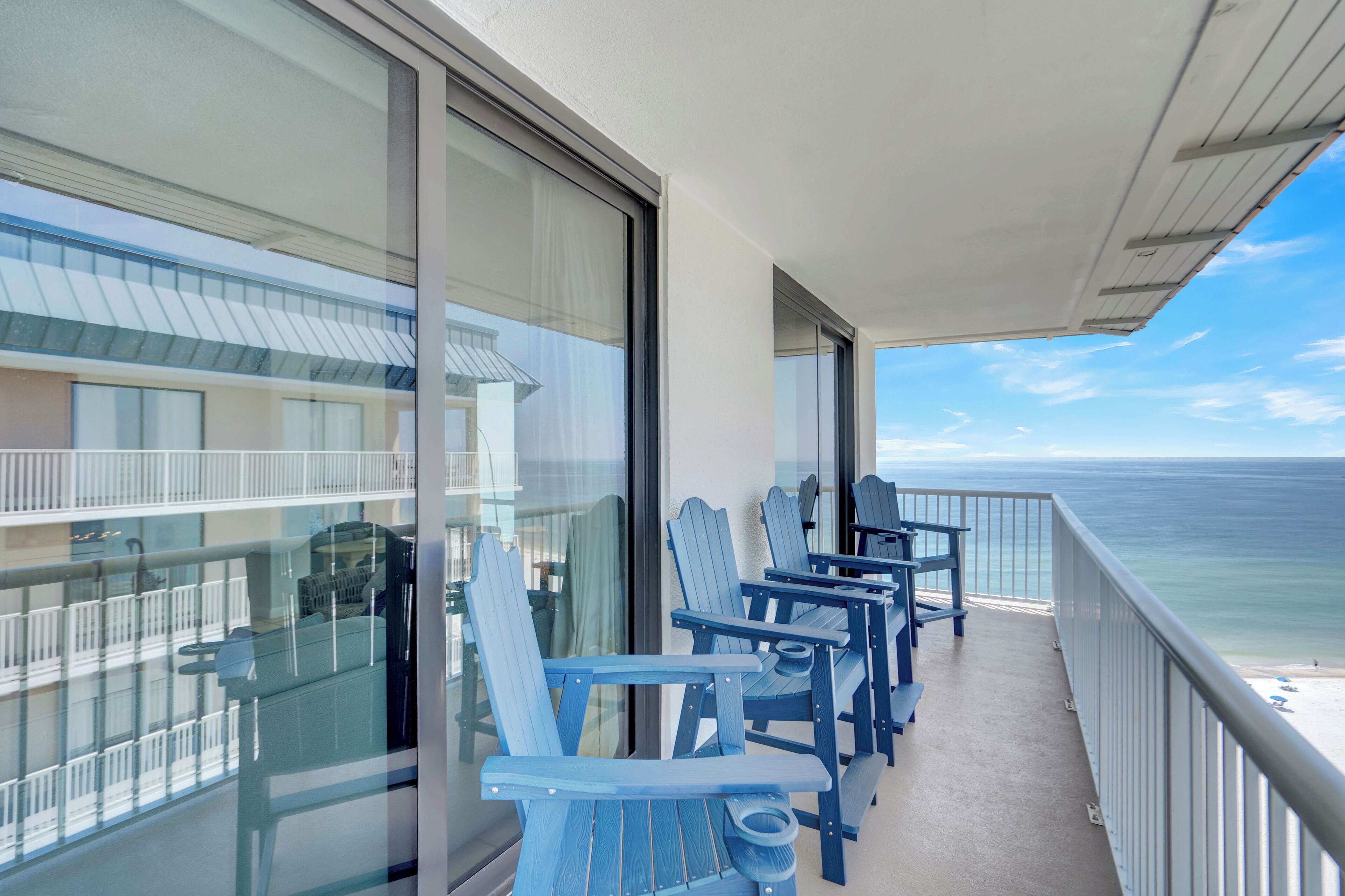 A balcony with blue Adirondack chairs, a glass railing, and a view of the ocean.