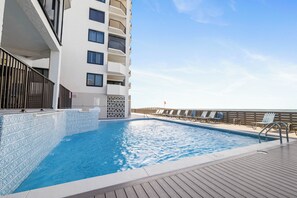 Une piscine avec une terrasse en bois, un bâtiment blanc doté de balcons et un ciel dégagé.