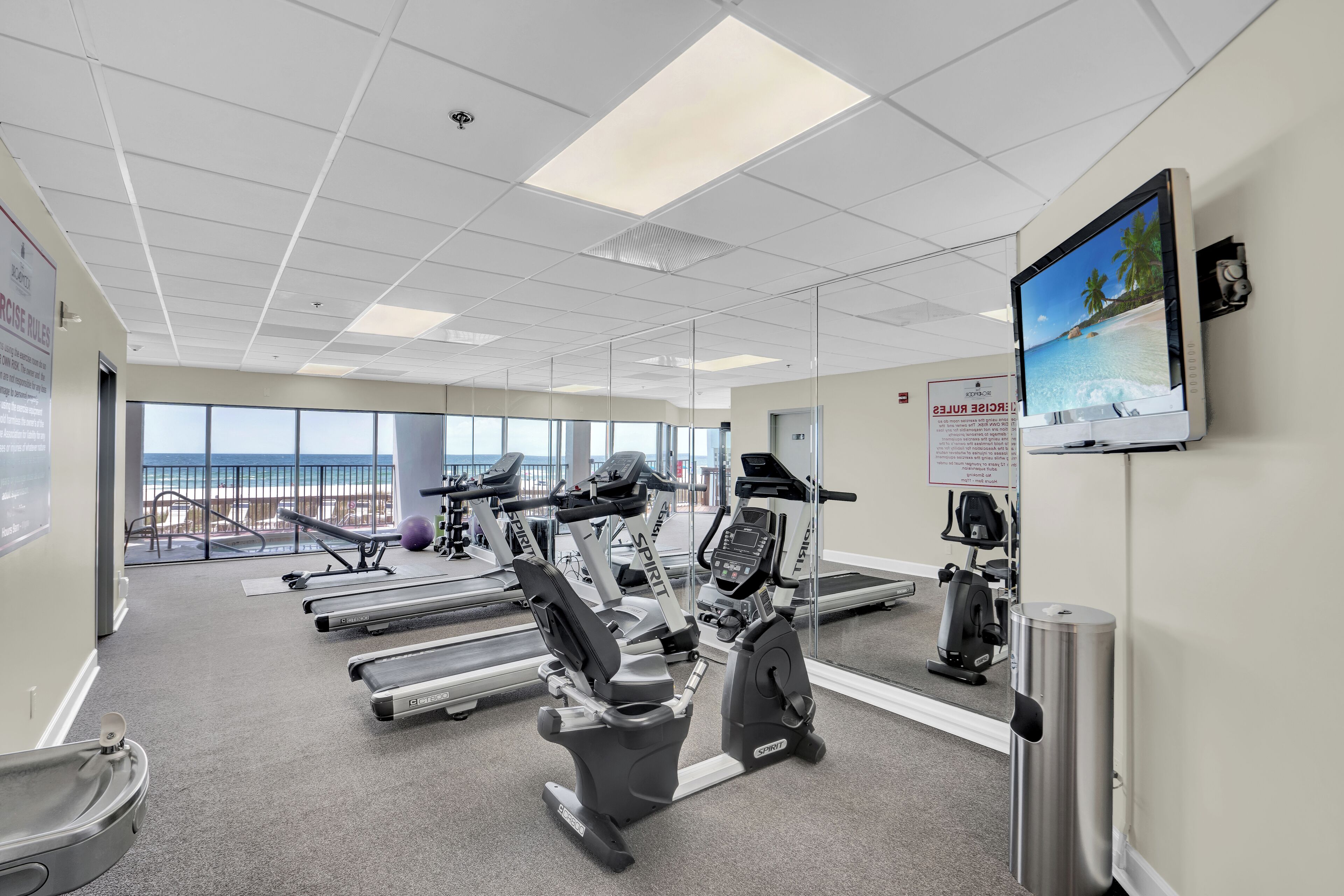 Ein Fitnessraum mit Cardiogeräten, einem großen Spiegel und einem Fernseher, der eine Strandansicht zeigt.