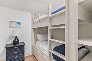 Een stapelbed met nachtkastje en lamp, een lijstfoto aan de muur, en een kleine dressoir met lades.
