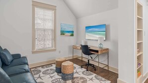 A room with a sloped ceiling, a desk with a computer, a black office chair, a blue sofa, two woven ottomans, a patterned rug, a window with blinds, and a shelf with books. - Golf Cart+4 Bikes! 5 Min to the Pool, 10 to Beach! 2 King Suites & Custom Bunks! ~ Baha Beach House at WaterSound West on 30A (Santa Rosa Beach)