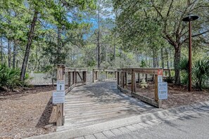 A wooden boardwalk leading to a beach, surrounded by trees. Signs indicate a bike path to the beach and a slow cycle speed limit of 1 MPH.