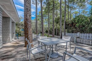 Een terras buiten met een metalen tafel en stoelen, omgeven door hoge bomen en een houten terras.