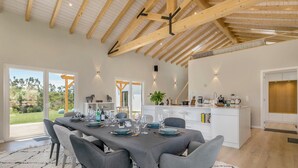 Un coin repas avec une longue table dressée pour six personnes, une cuisine en arrière-plan et une vue sur l’extérieur.