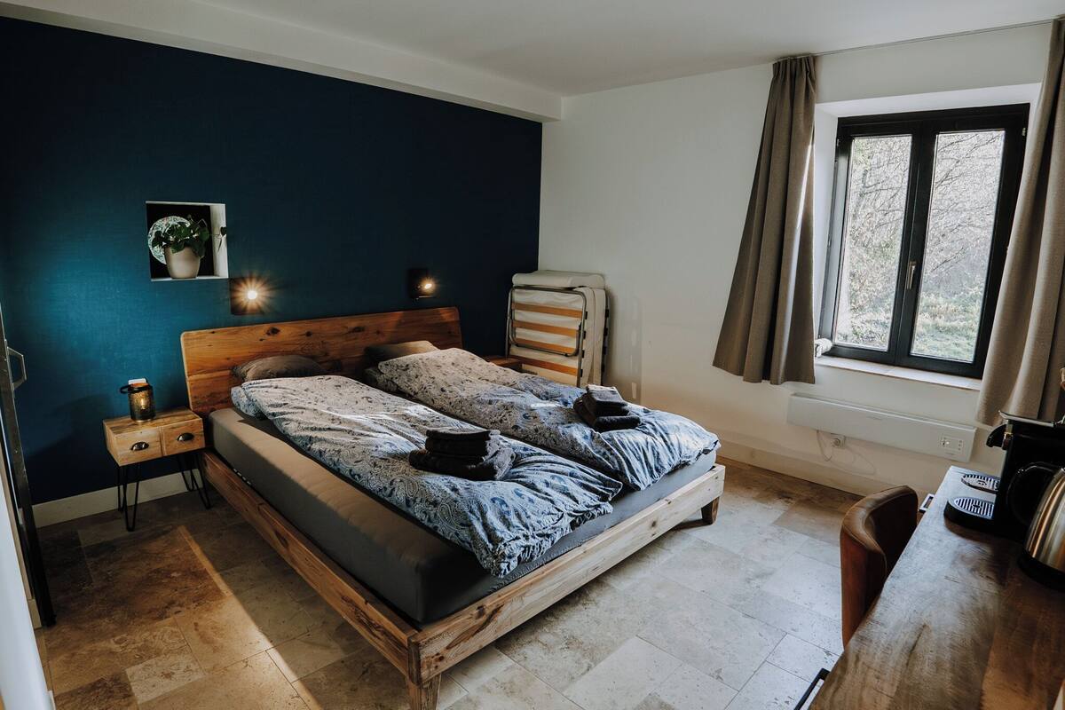 Une chambre avec un lit en bois, une table de chevet, une petite table, une fenêtre avec des rideaux et une plante en pot accrochée au mur.