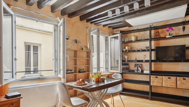 A dining area with a wooden table, white chairs, and a view of the outside. There are shelves with decorative items and a television on the wall.