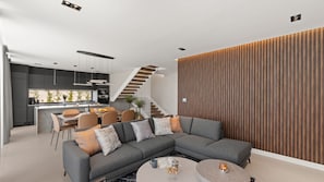 Un soggiorno moderno con un divano ad angolo grigio, una zona pranzo con tavolo e sedie bianche, una cucina con mobili scuri e una parete in listelli di legno.