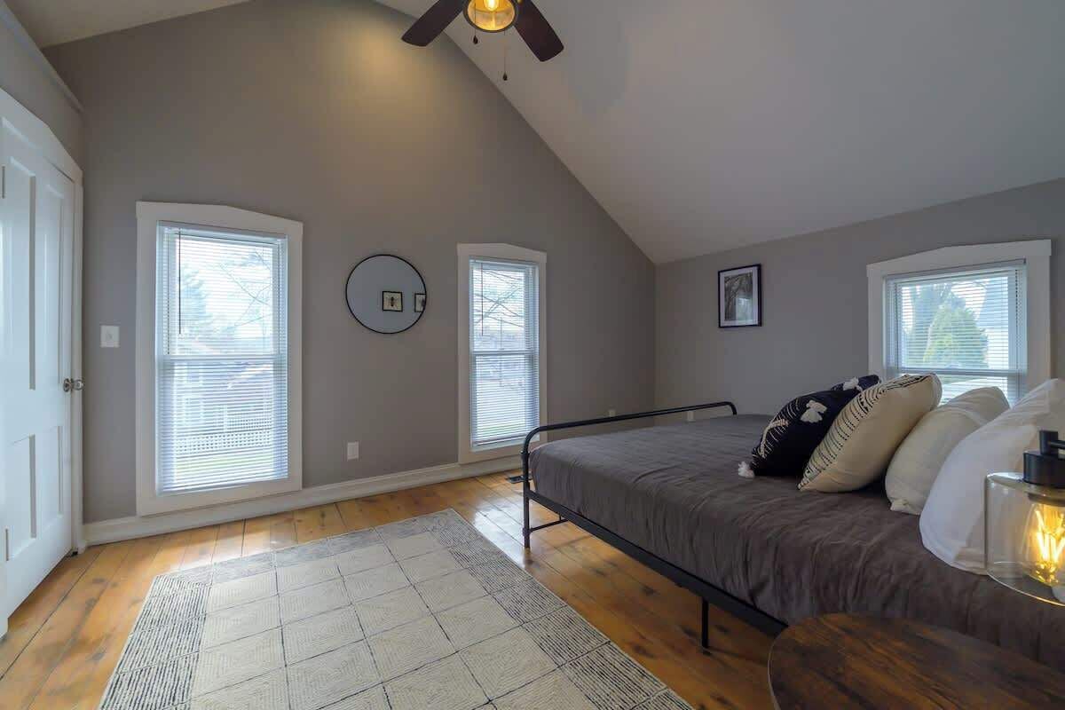 A bedroom with a sloped ceiling, two windows with blinds, a bed with a metal frame, a rug, and a ceiling fan.