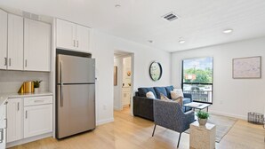 A modern kitchen with white cabinets and a stainless steel refrigerator. There is a grey sofa and a small table with a plant in the living area. - MLK #104 xxxxxxxxxxx (Austin)