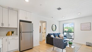 Una cocina moderna con armarios blancos y un refrigerador de acero inoxidable. Hay un sofá gris y una pequeña mesa con una planta en la sala.