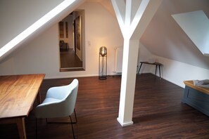A modern loft bedroom with a sloped ceiling, a wooden dining table, a light blue chair, a floor lamp, and a small table with a towel.