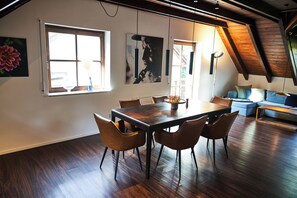 A dining area with a wooden table and chairs, large windows, and a loft-style ceiling.