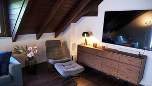 A modern living room with a high ceiling, wooden beams, a grey armchair, a small ottoman, a wooden cabinet, a TV, and a vase with flowers. - Romantic rooftop apartment (Windach)