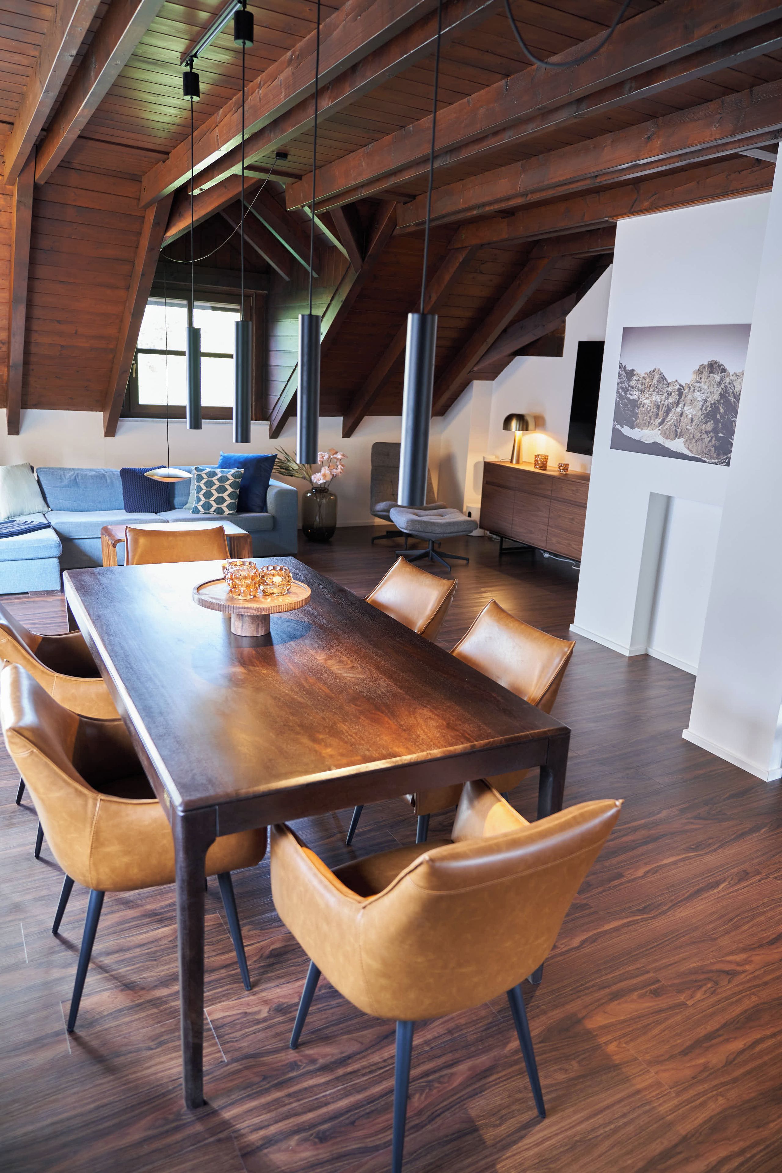 A spacious loft with a high ceiling, exposed wooden beams, a dining area with a long table and leather chairs, a cozy seating area with a sofa and cushions, and a large window allowing natural light.