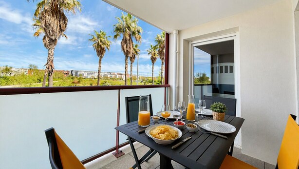A balcony with a table set for breakfast, overlooking palm trees and a distant building.