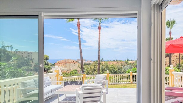 Un balcon avec vue sur la mer, des meubles d’extérieur blancs et un parasol rouge.