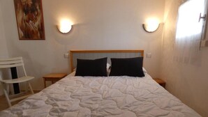 Ein ordentlich bezogenes Bett mit weißer Bettdecke und zwei schwarzen Kopfkissen, ein weißer Sessel und eine Wandleuchte.