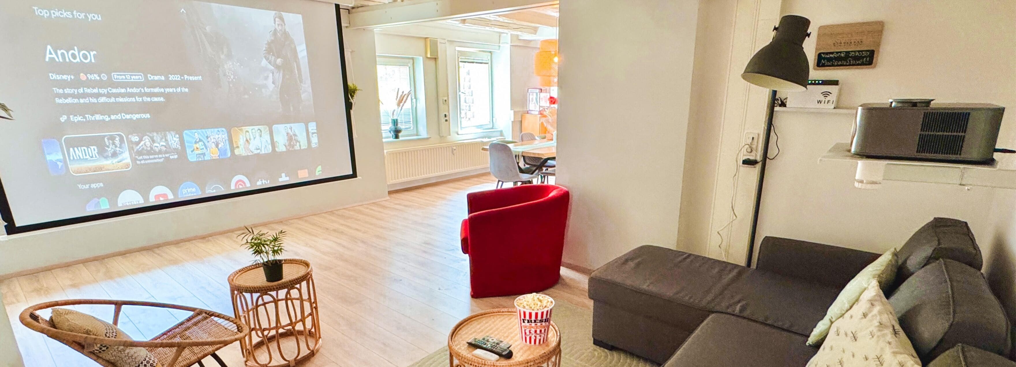 Un salon avec un grand écran diffusant un film, un fauteuil rouge, un canapé gris et une petite table avec un bol de pop-corn.