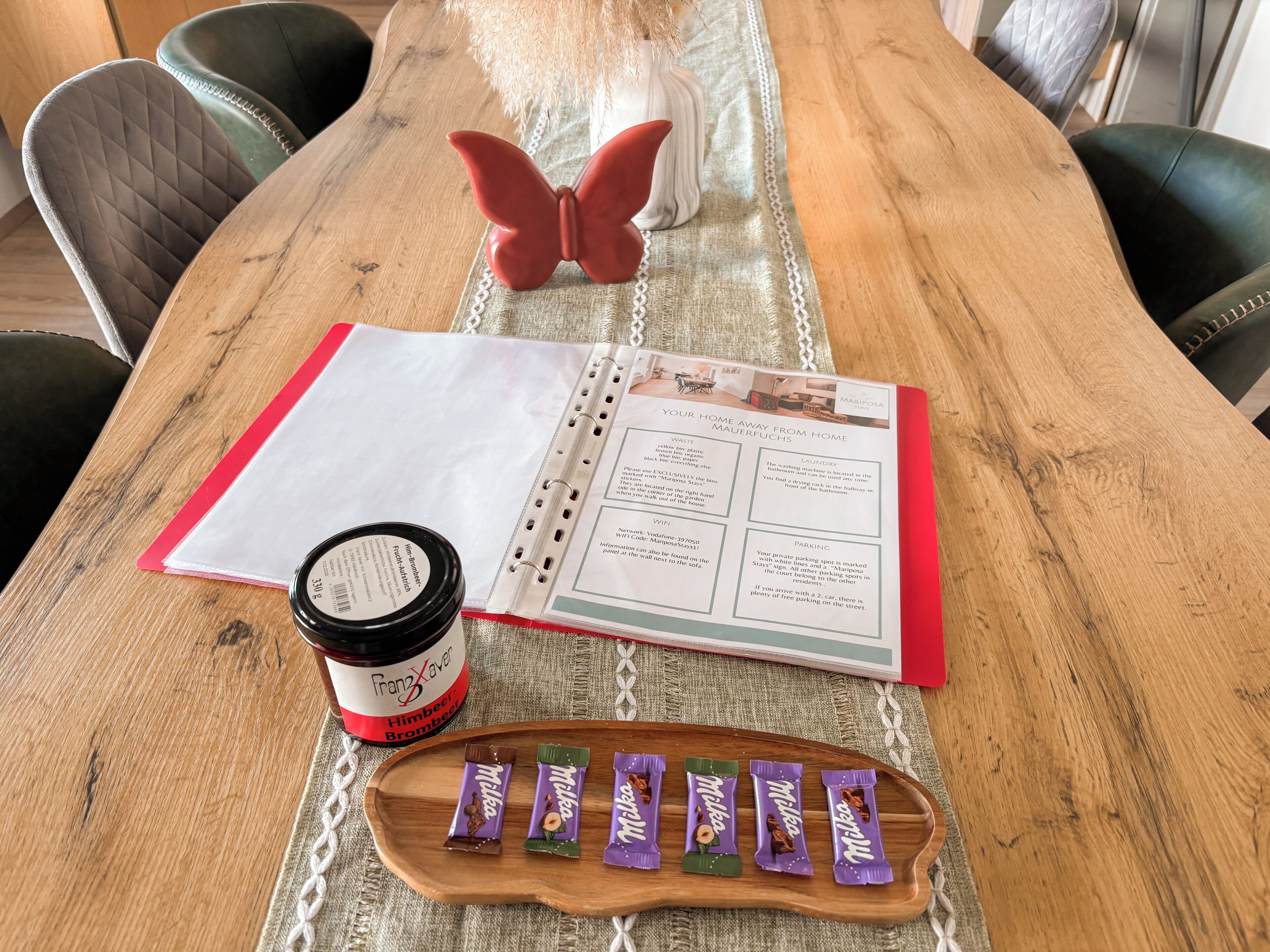 Ein Holztisch mit einem roten und weißen Planer, einer Marmeladenglas und einer Schale mit Milka Schokoriegeln.
