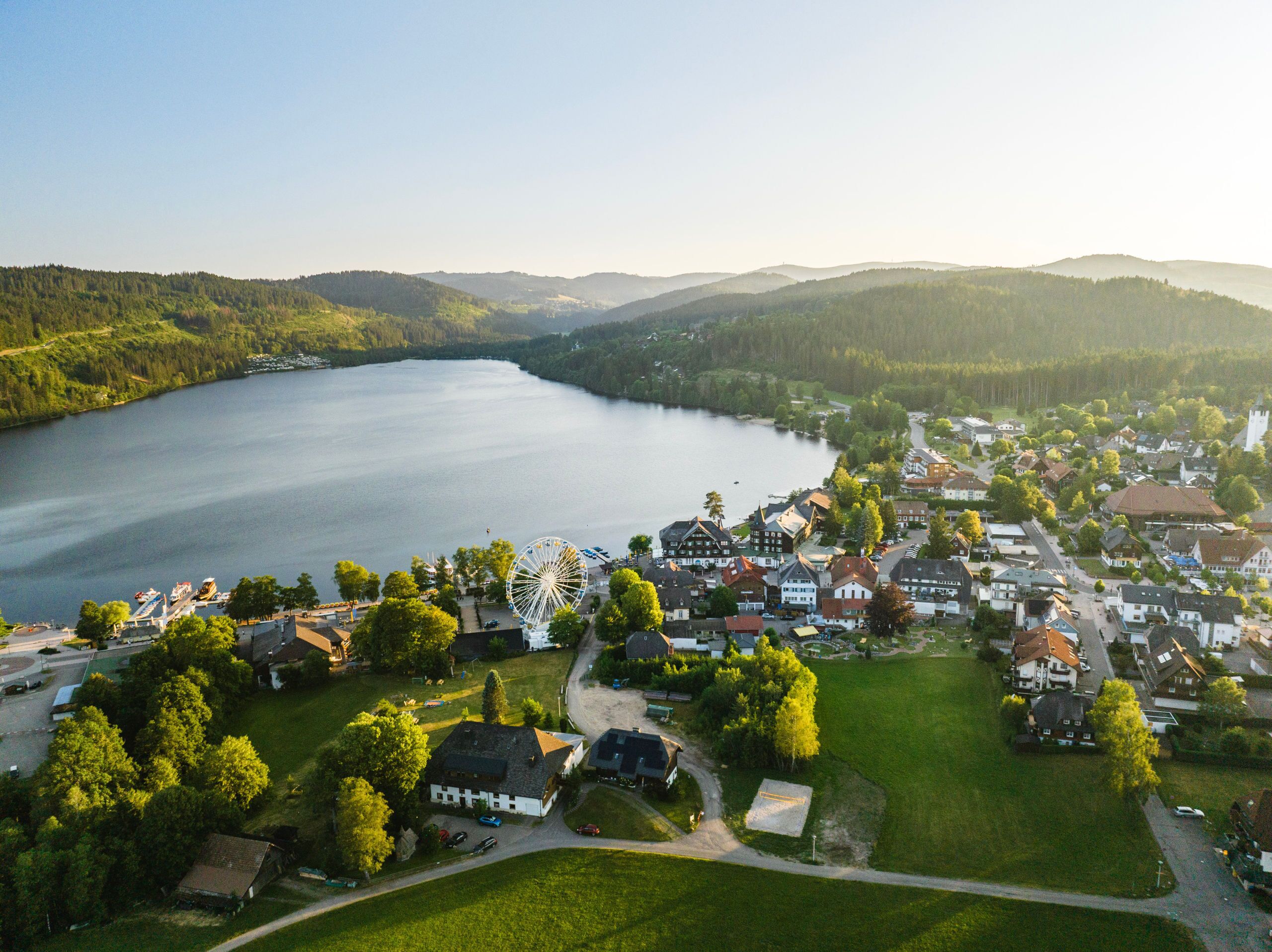 Vista aérea de un pueblo junto al lago, con una noria destacada, rodeado de edificios residenciales y exuberante vegetación.