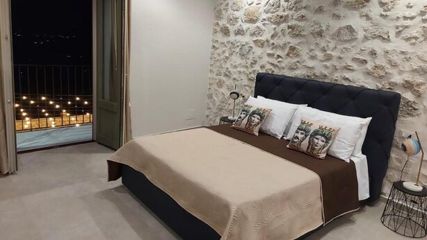 A bedroom with a stone wall, a bed with a dark headboard, and a view of a balcony with lights. - Masseria Torre Saracena (Favara)