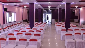 A conference room with rows of white chairs with red sashes, purple pillars, and a ceiling with recessed lighting. - Hotel Roop Shree Inn By GRB (Patna)