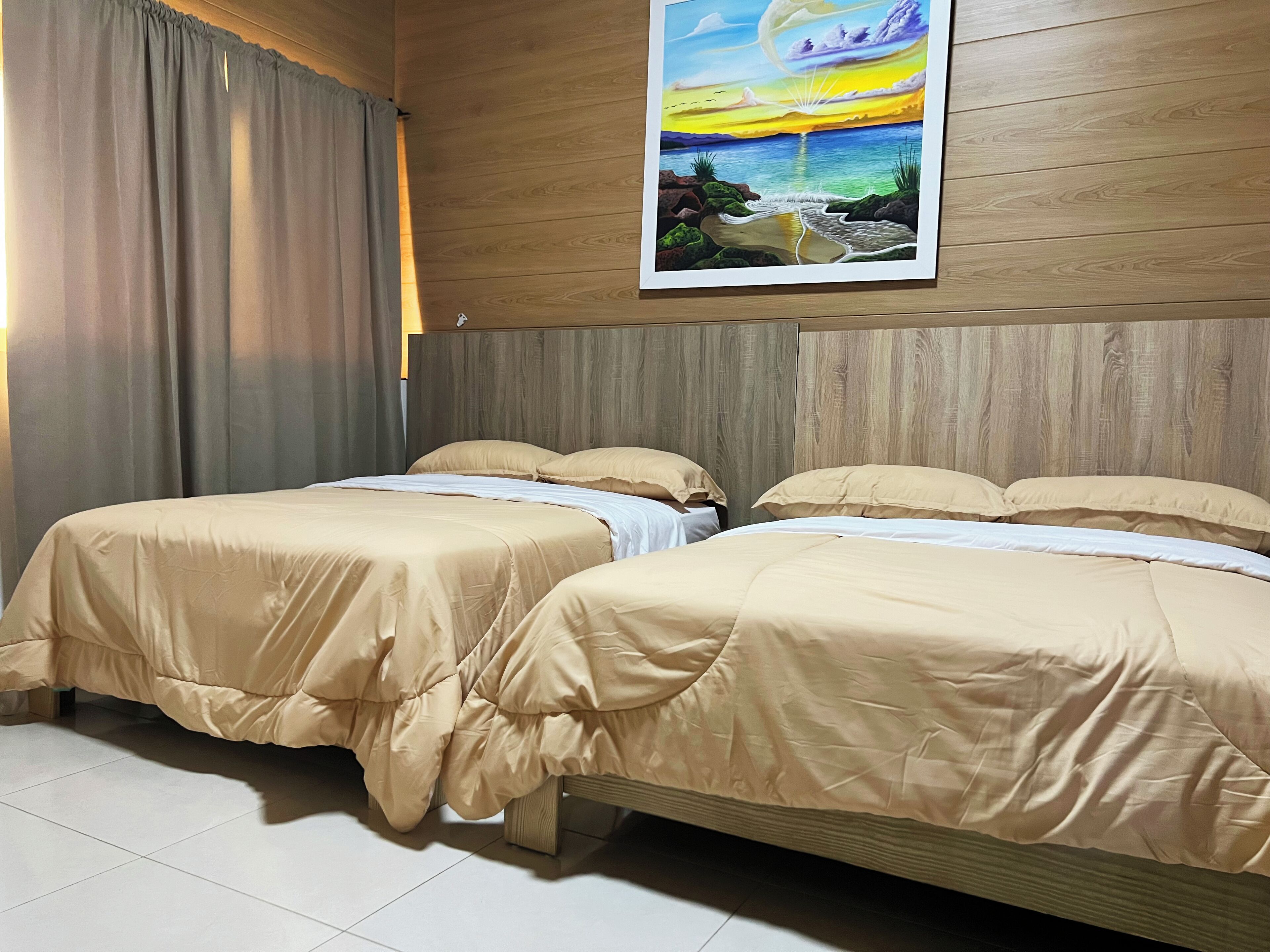 Ein Hotelzimmer mit zwei Betten, einem Holzkopfteil, beiger Bettwäsche, einem Sonnenuntergangsgemälde und einem Vorhang am Fenster.