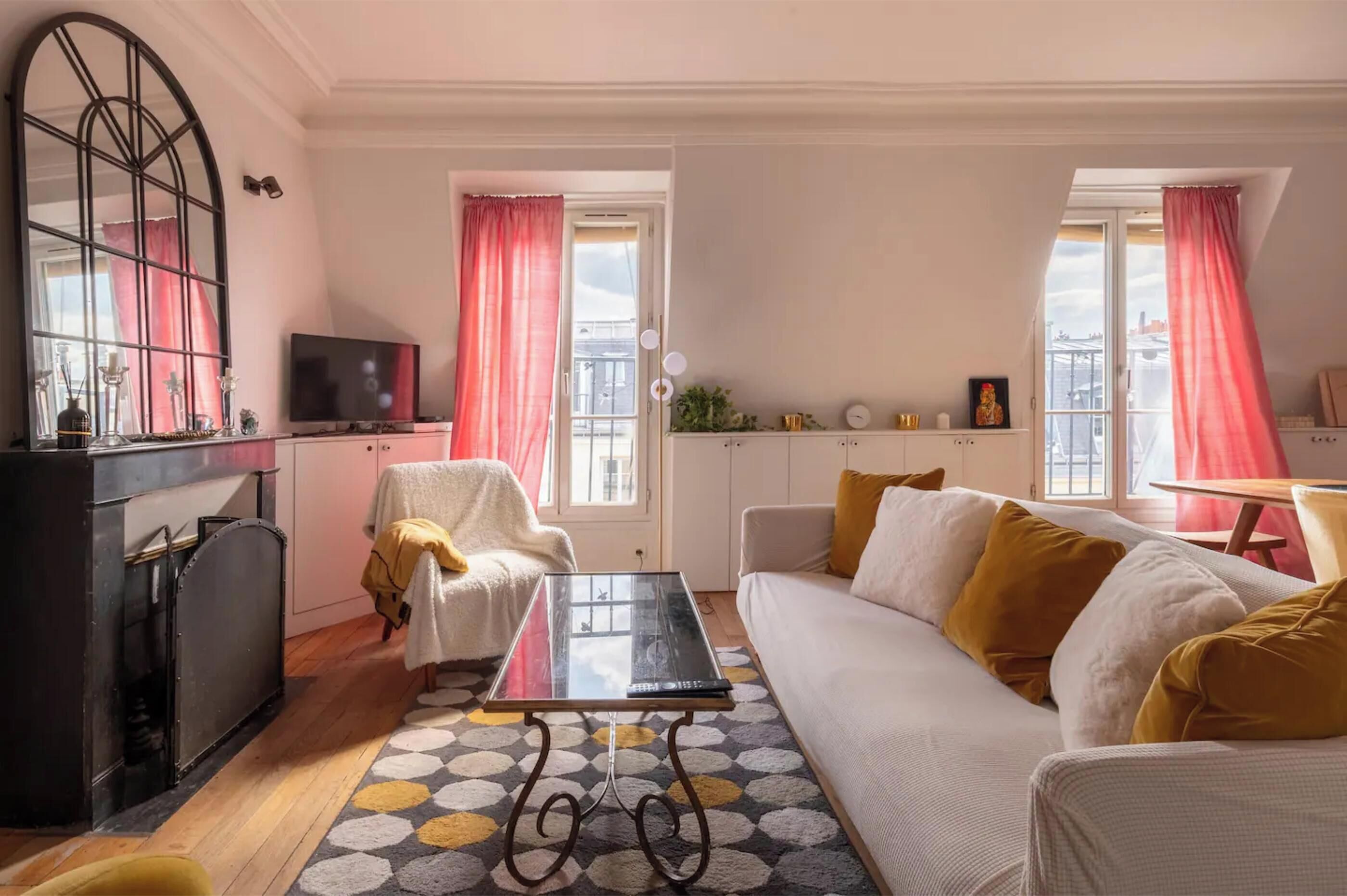 A living room with a sofa, a coffee table, a TV, and a cabinet. There are windows with curtains, a patterned rug, and a small table with a chair.