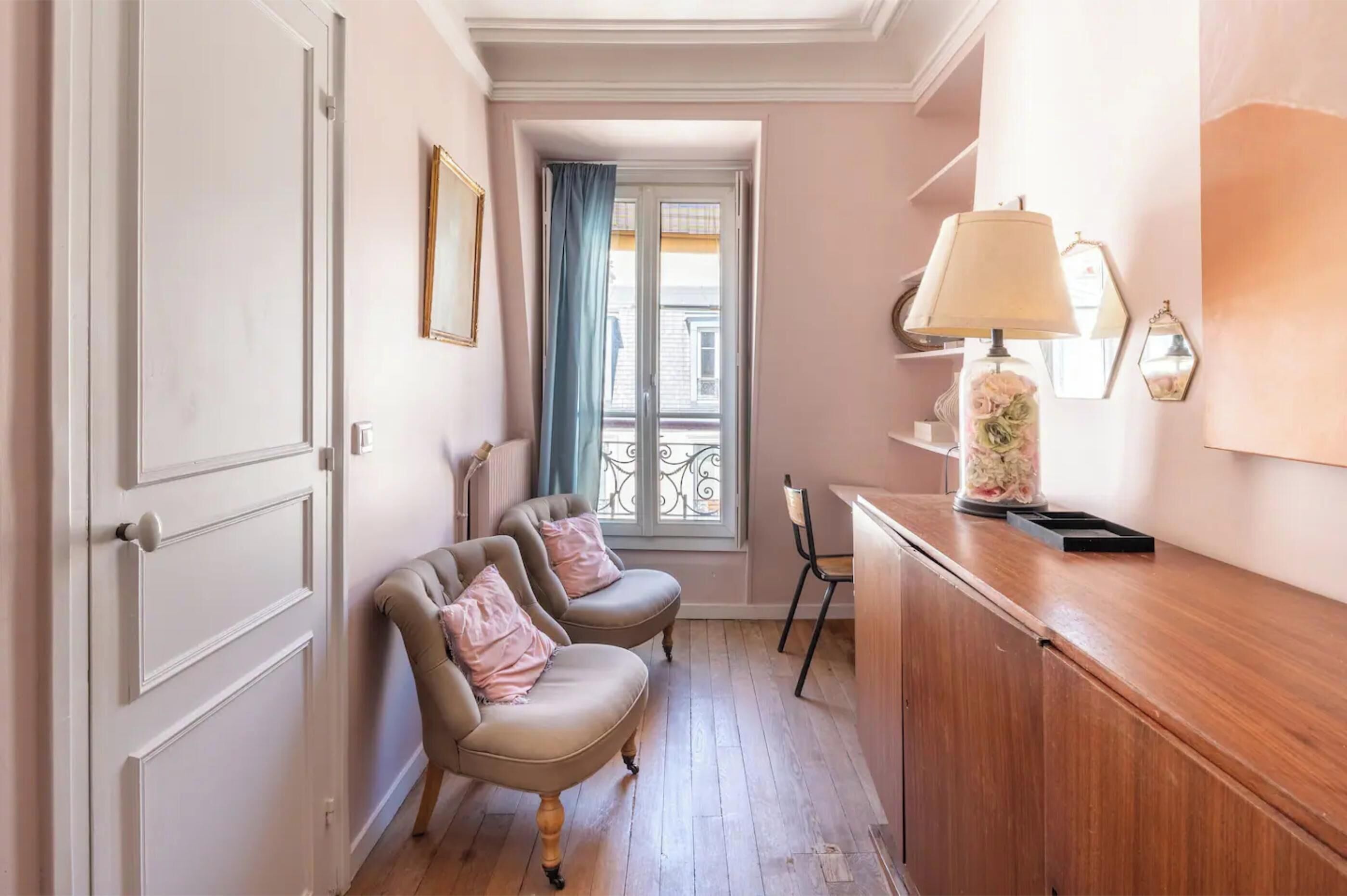 A hallway with two upholstered chairs, a wooden cabinet, a lamp, and a window with a view of buildings.