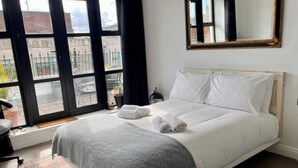 A neatly made bed with white linens and a grey throw blanket, a small bedside table with a lamp, and a large window with a view of the outside. - 2 Bed City Centre Penthouse with Cathedral Views (Dublin)