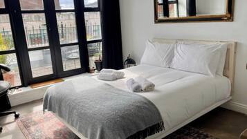 A neatly made bed with white linens and a grey throw blanket, a small bedside table with a lamp, and a large window with a view of the outside.