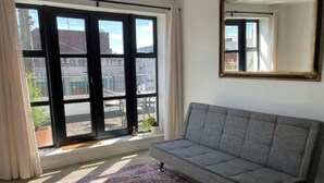 Une pièce avec un canapé gris, un tapis à motifs, une grande fenêtre aux montures noires et un miroir au mur.