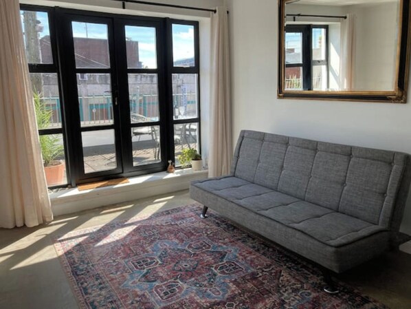 A room with a grey sofa, a patterned rug, a large window with a view of buildings, and a mirror on the wall.