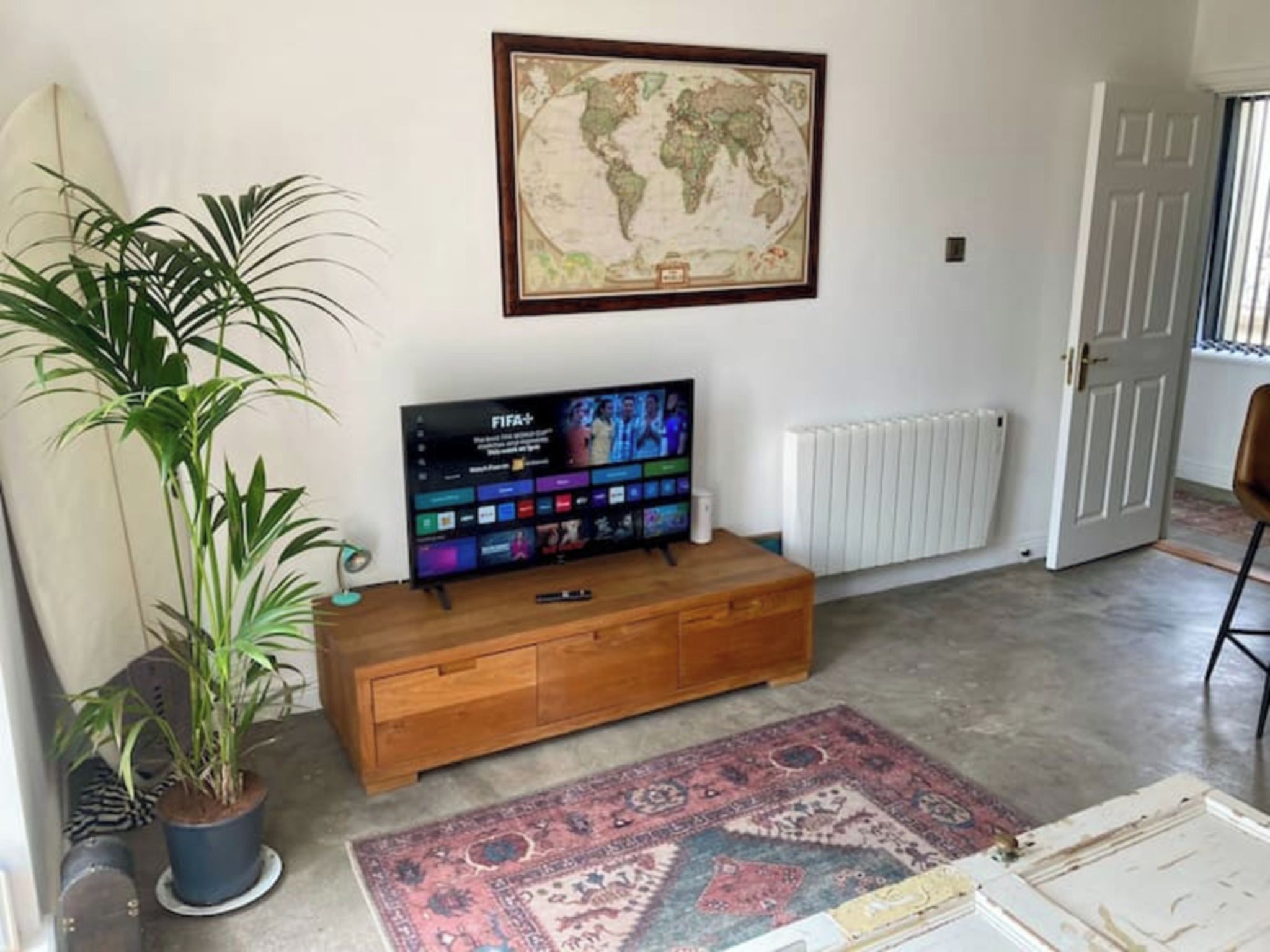 A living room with a wooden TV stand, a flat-screen TV, a world map on the wall, a potted plant, and a patterned rug.