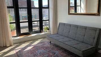 A room with a grey sofa, a patterned rug, a large window with a view of buildings, and a mirror on the wall.